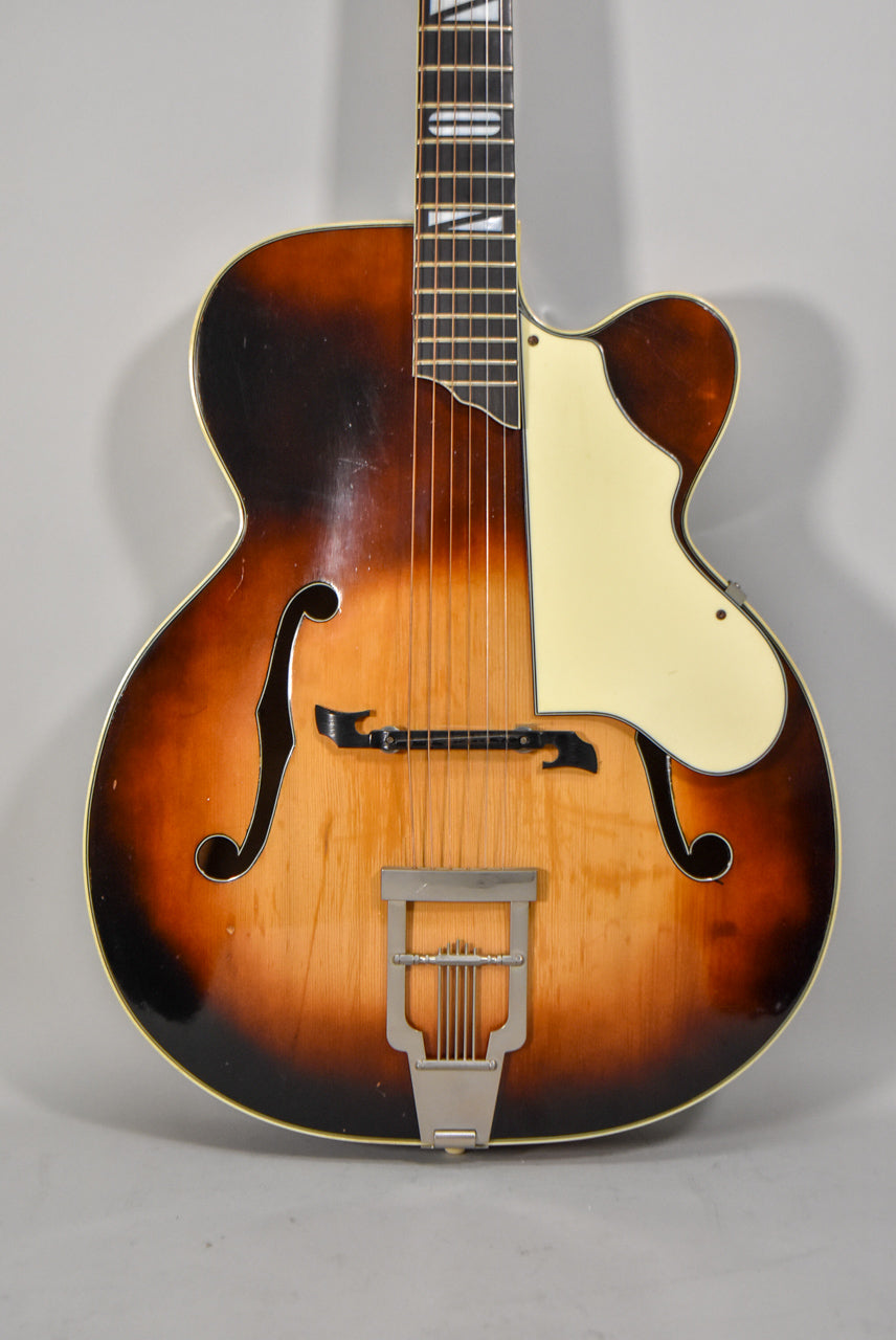 Full front view of a sunburst archtop guitar with a cream pickguard, f-holes, and a trapeze tailpiece.