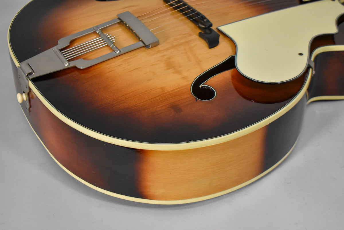 Close-up of the sunburst archtop guitar's lower bout, showing the F-hole, pickguard, and trapeze tailpiece with some wear.