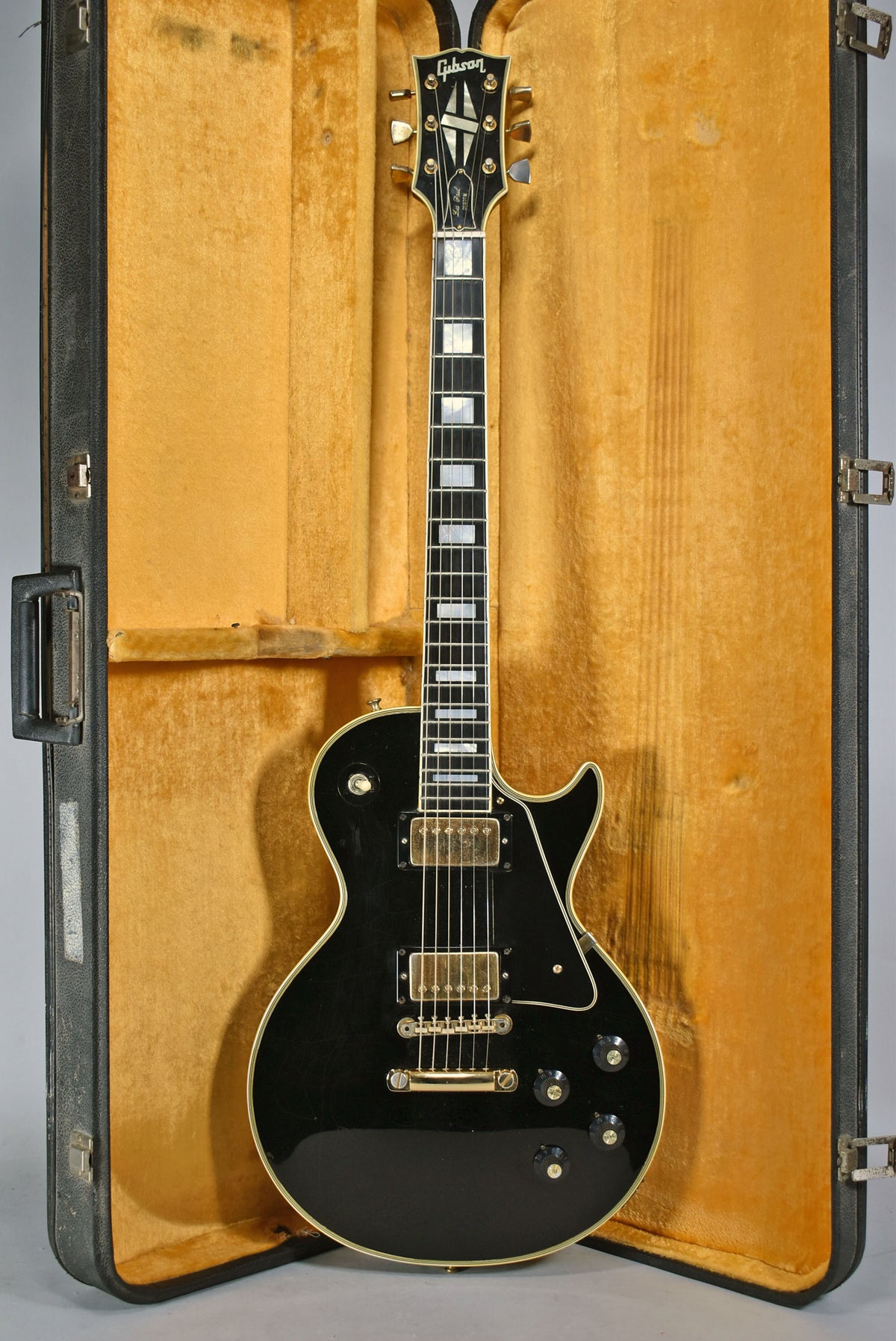 Full-length view of a black electric guitar with gold hardware and white binding, resting in an open, yellow-lined hard case.