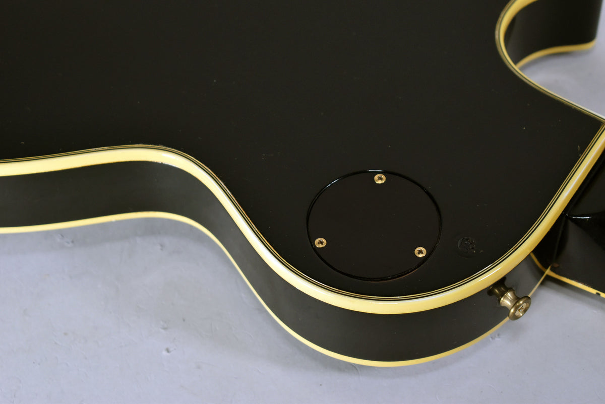 The black guitar body's lower bout, showing the control cavity cover with three gold screws and a gold strap button.