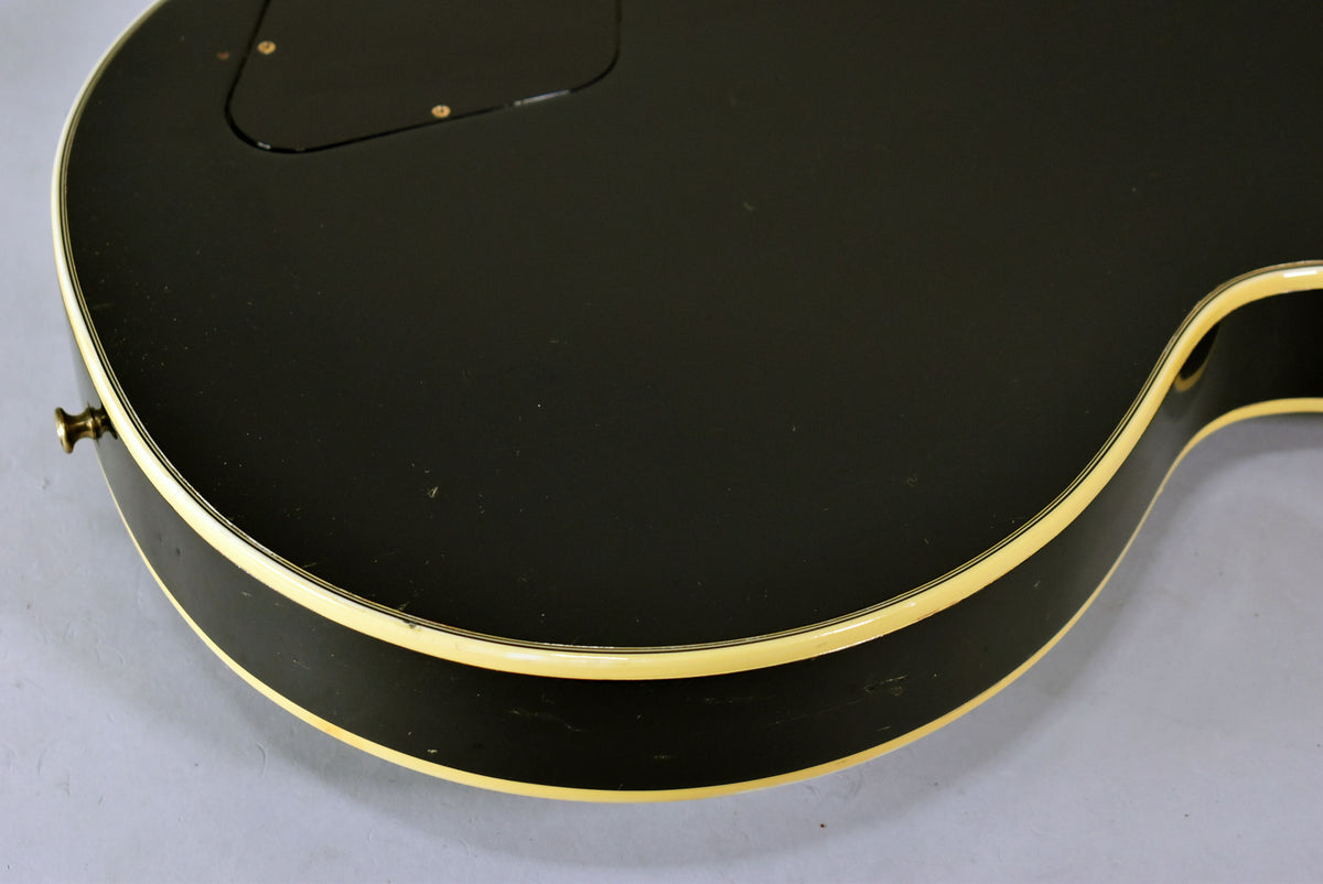 Close-up of the guitar's black body with cream binding, showing light wear and a control cavity cover with two gold screws.