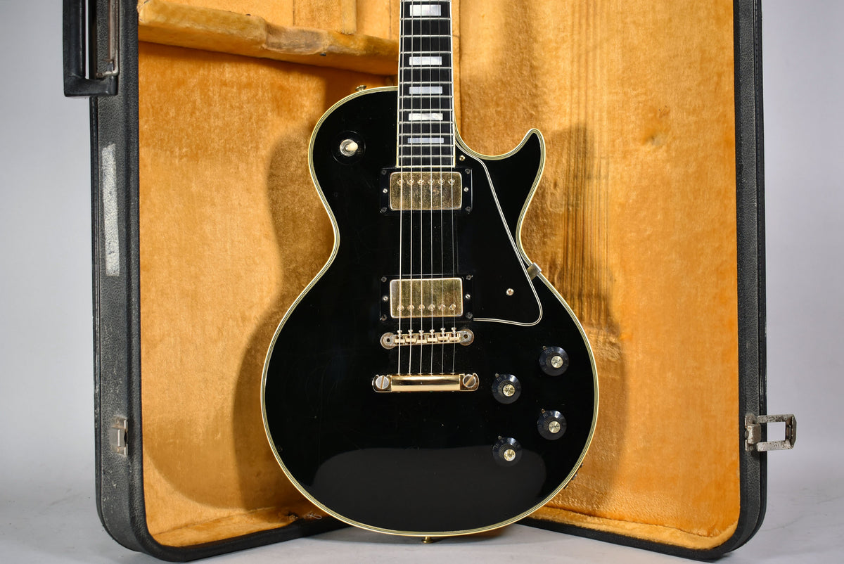 Black electric guitar with gold hardware, visible checking on the body, and rectangular fretboard inlays, resting in a tan-lined case.