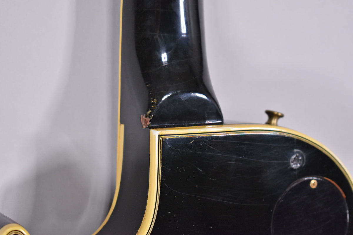 Close-up of the guitar's black body and neck joint, showing finish wear and a chip on the neck heel, with a gold strap button.