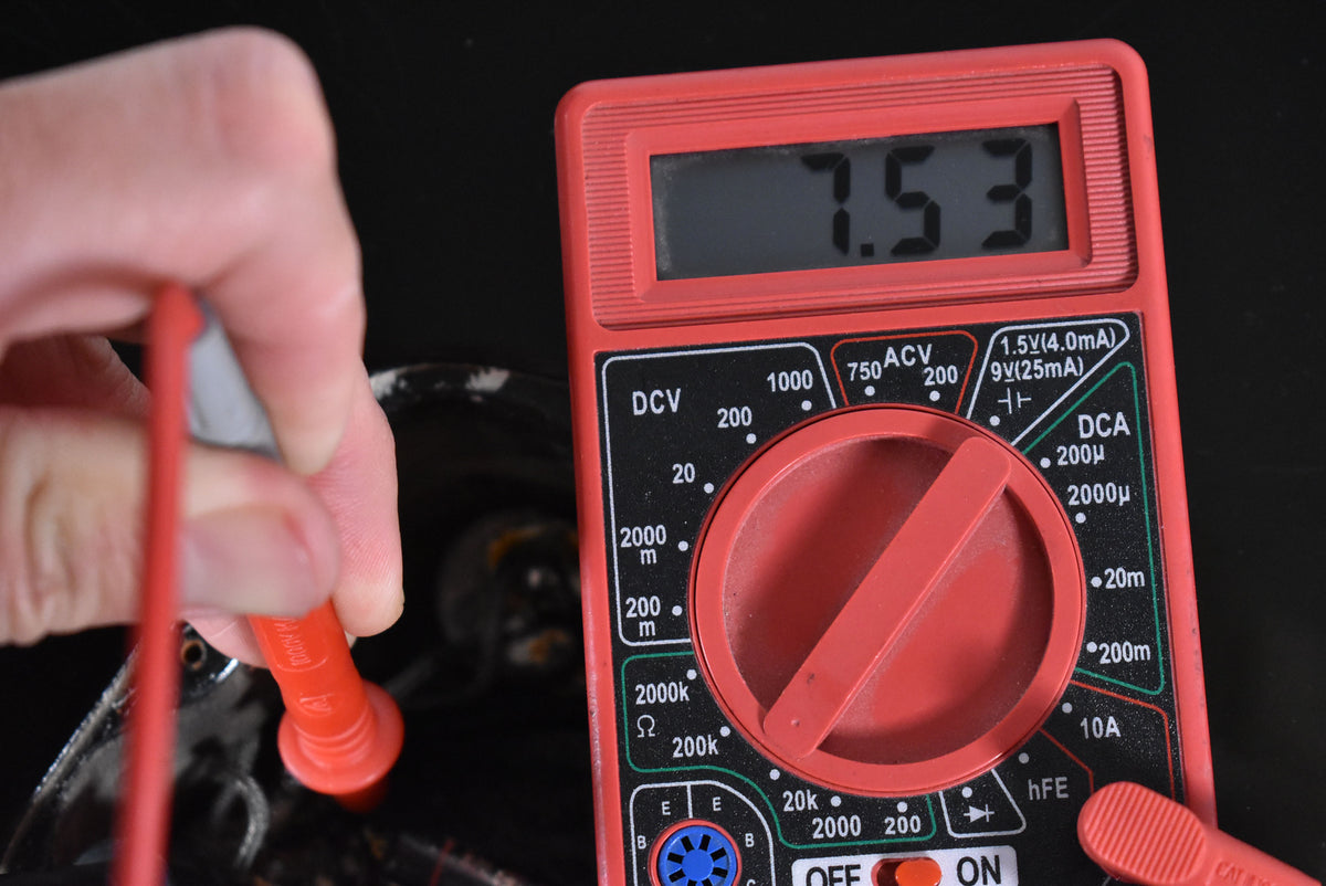 A red multimeter shows a reading of 7.53, with a hand holding a red probe touching a dark, worn part of the guitar.