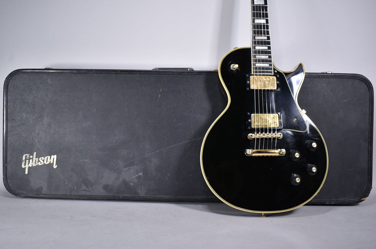 Black electric guitar with gold hardware and white binding, standing upright in front of its worn black hard case.