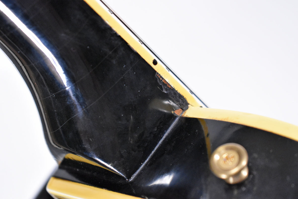 Close-up of the black guitar's neck heel, showing finish checking, binding separation, and a chip revealing the wood beneath.