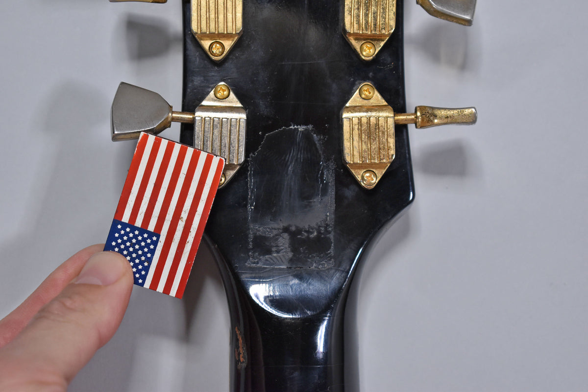 Close-up of the black headstock's rear, showing four gold tuners, wear, and a hand holding a small US flag sticker.