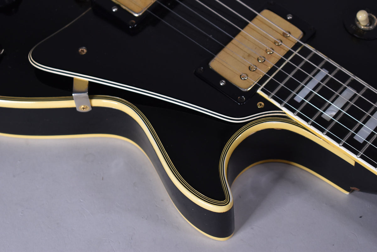 Close-up of the black guitar body's lower bout, showing cream binding, gold humbuckers, and rectangular fretboard inlays.
