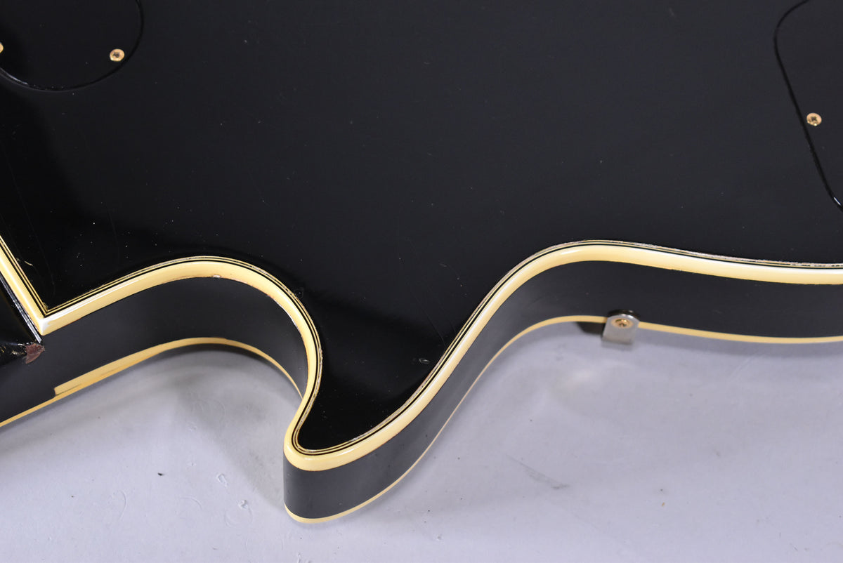 The lower bout of a black electric guitar, showing cream binding with thin black stripes, a strap button, and some finish wear.