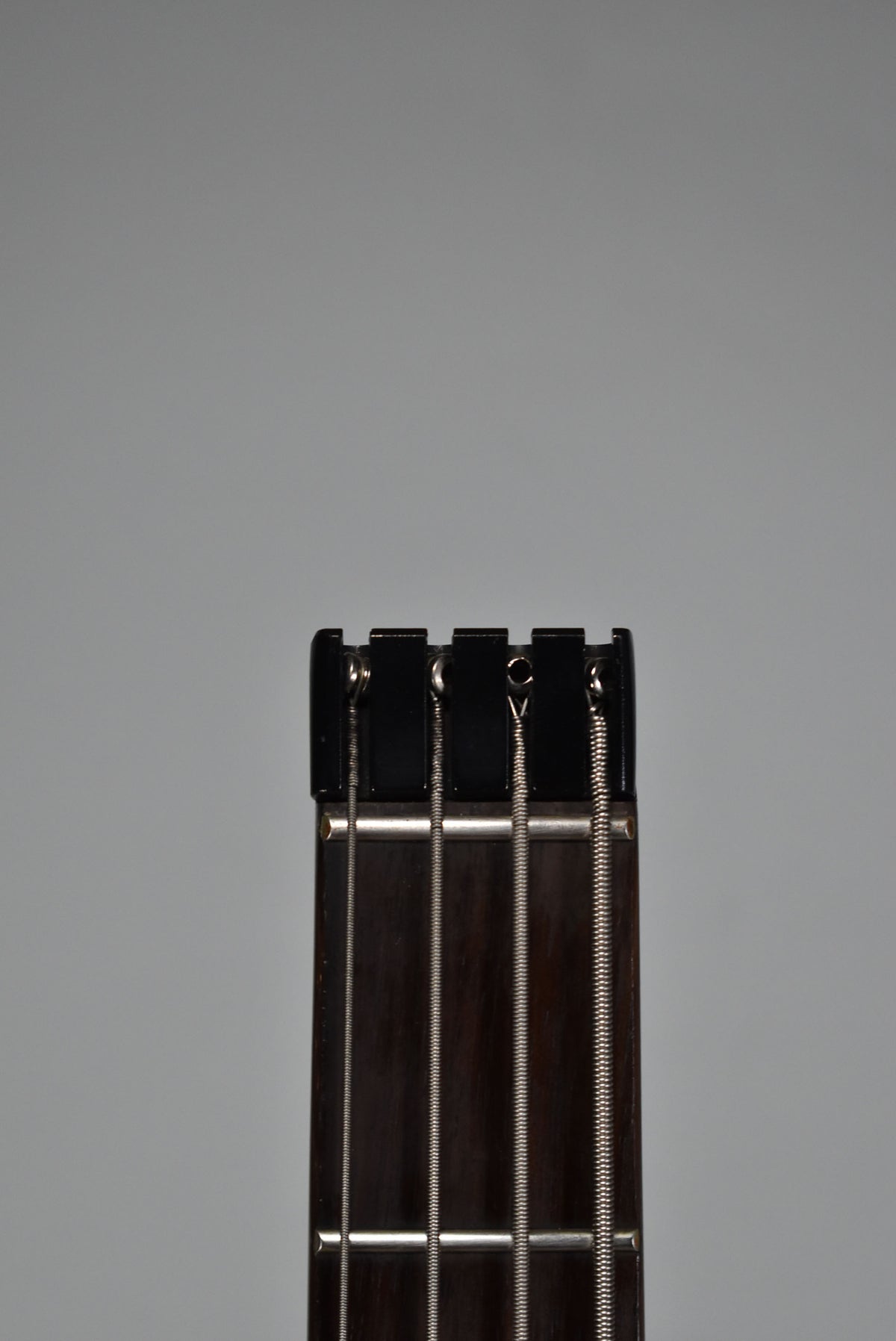Close-up of the bass guitar's dark fretboard and black headpiece, showing four silver strings and frets against a grey background.