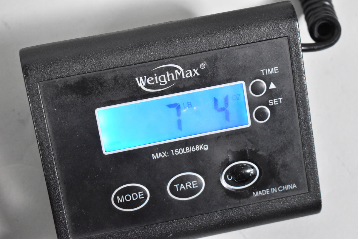 Close-up of a black digital scale displaying "7 LB. 4 OZ." on a blue backlit screen, with "WeighMax" and "MADE IN CHINA" visible.