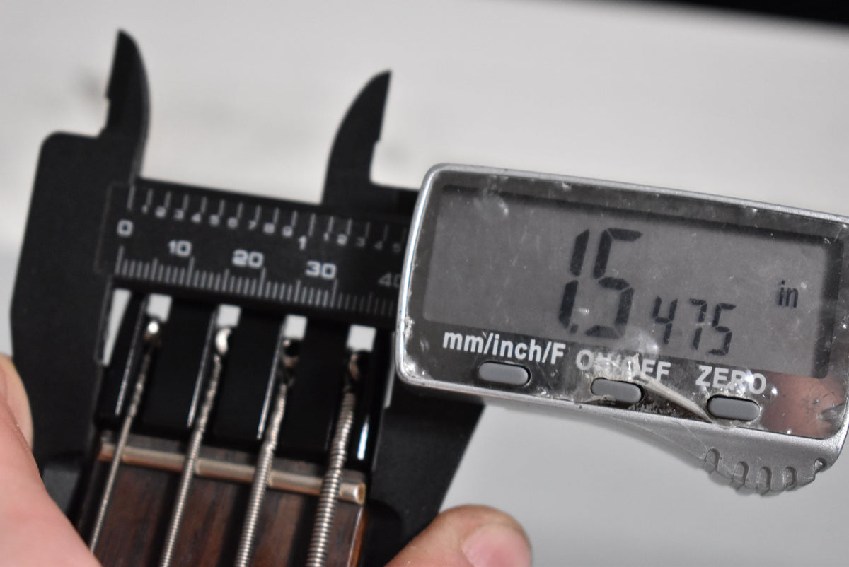 A close-up of a digital caliper measuring the nut width of the bass, showing a reading of 1.5475 inches.