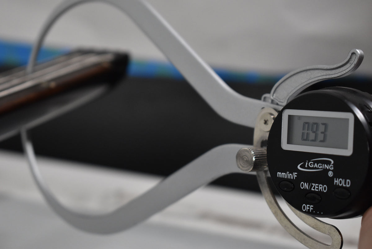 A digital caliper measures the bass's dark fretboard, showing "0.93" on its screen, with the strings blurred in the background.