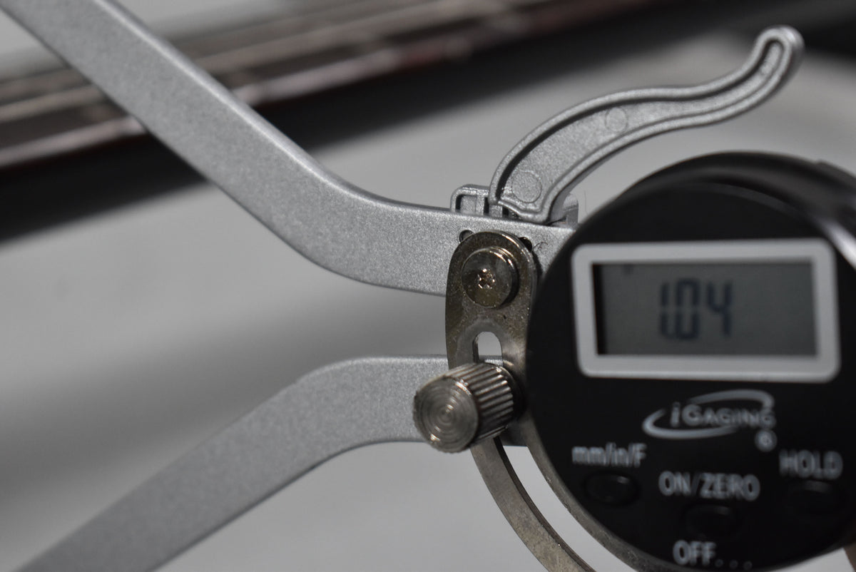 Close-up of a digital fret height gauge clamped onto a fret, displaying "1.04" on its screen.