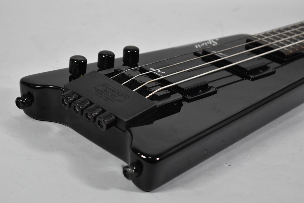 Close-up of the glossy black headless bass body, showing the tuning system, two humbucker pickups, and four strings.