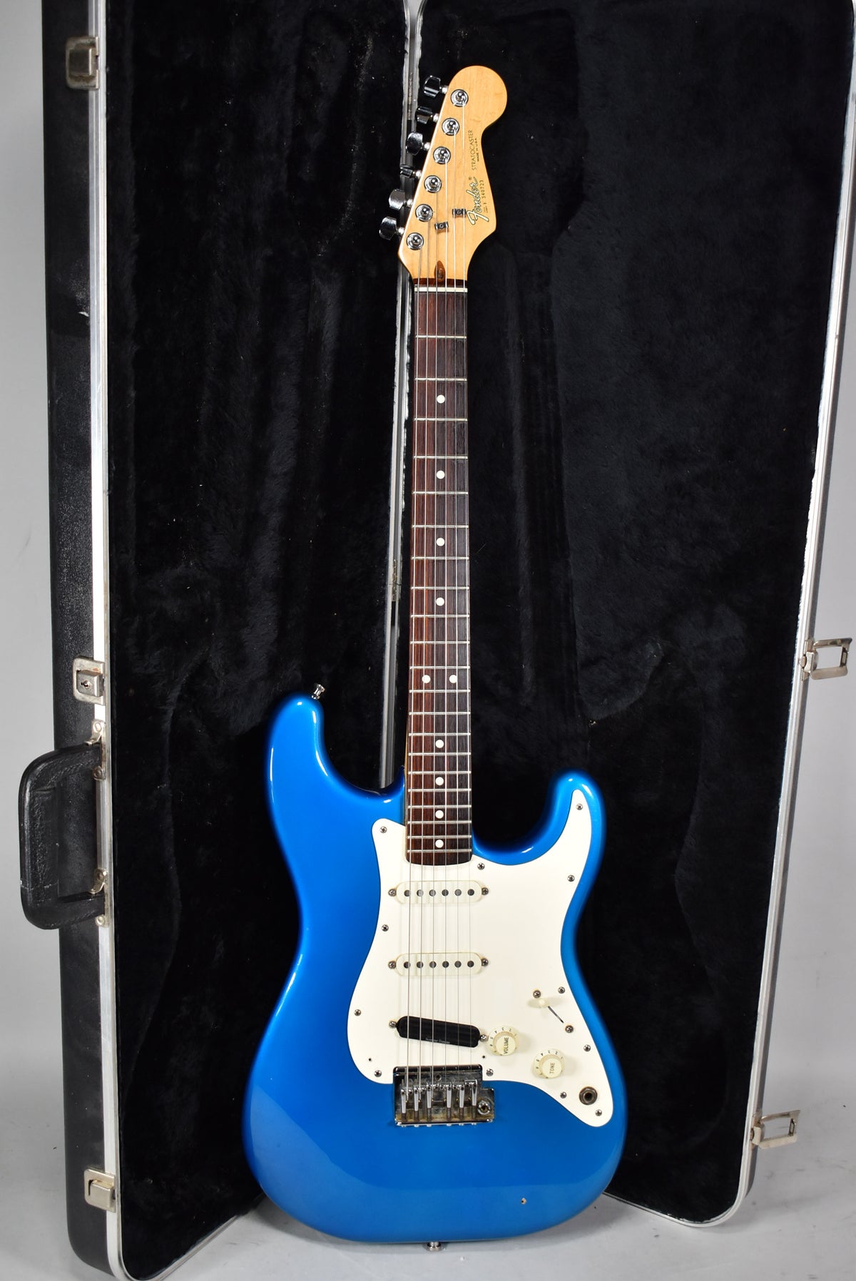 A full-length view of a glossy blue electric guitar with a white pickguard, three single-coil pickups, and a maple neck, resting in its open black hard case.