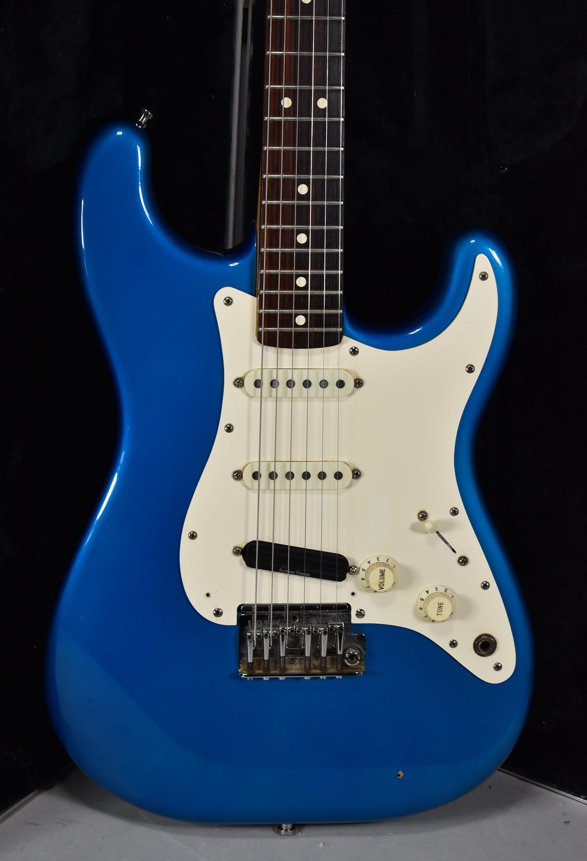 Blue electric guitar, angled from above, showing the body, white pickguard with three pickups, and part of the fretboard.