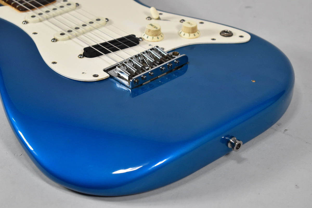 Close-up of a blue electric guitar body, showing the chrome bridge, white pickguard, cream knobs, and a small chip in the finish.