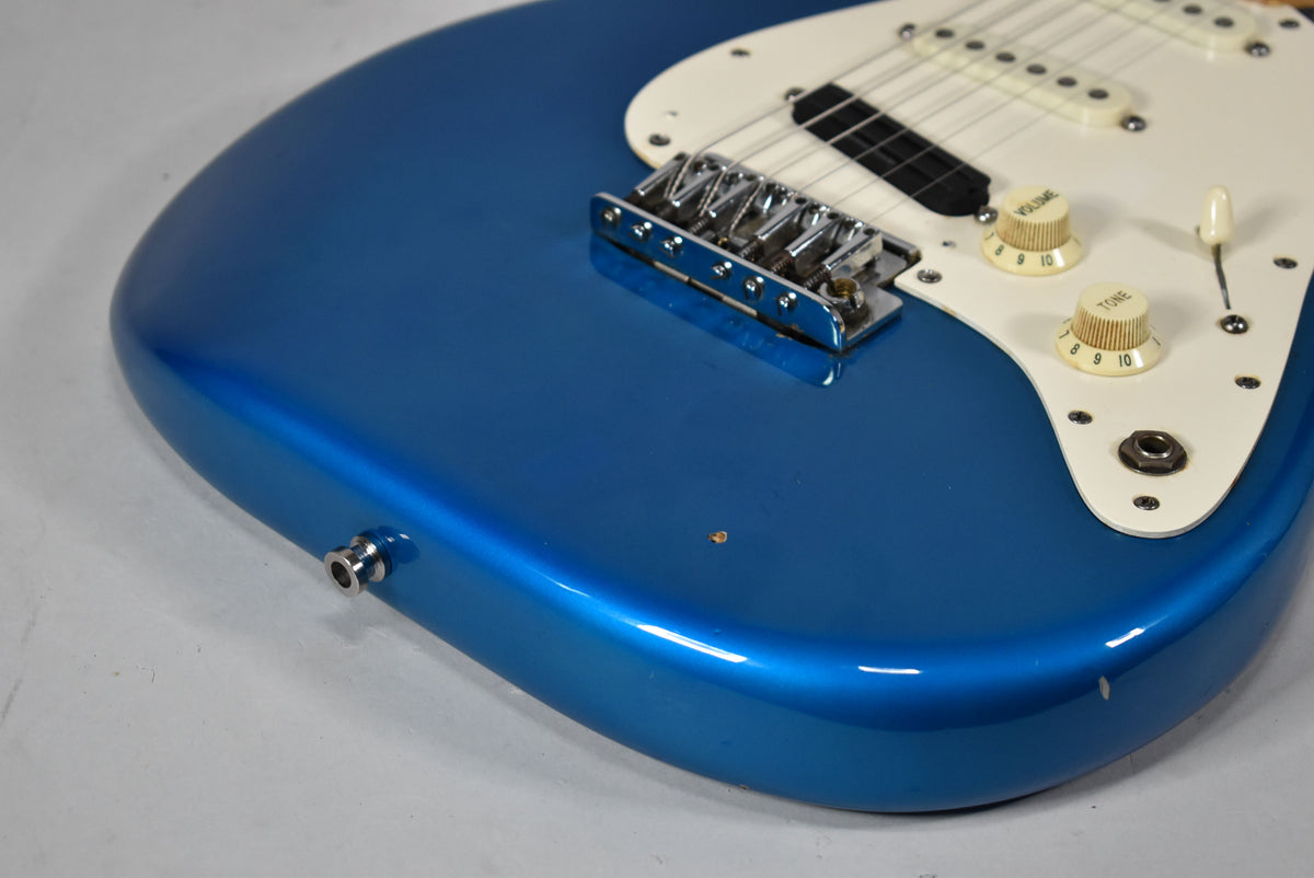 Close-up of the blue guitar body, showing the chrome bridge, white pickguard with cream knobs, and a small chip near the input jack.