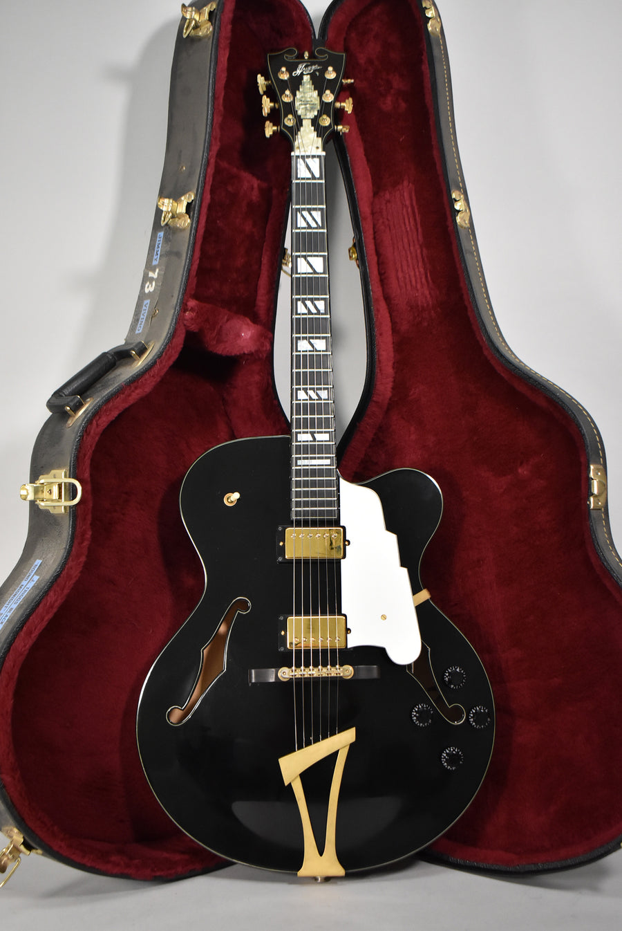 Full-front view of a glossy black archtop guitar with gold hardware, white pickguard, and trapezoid fretboard inlays, resting in its open red-lined case.