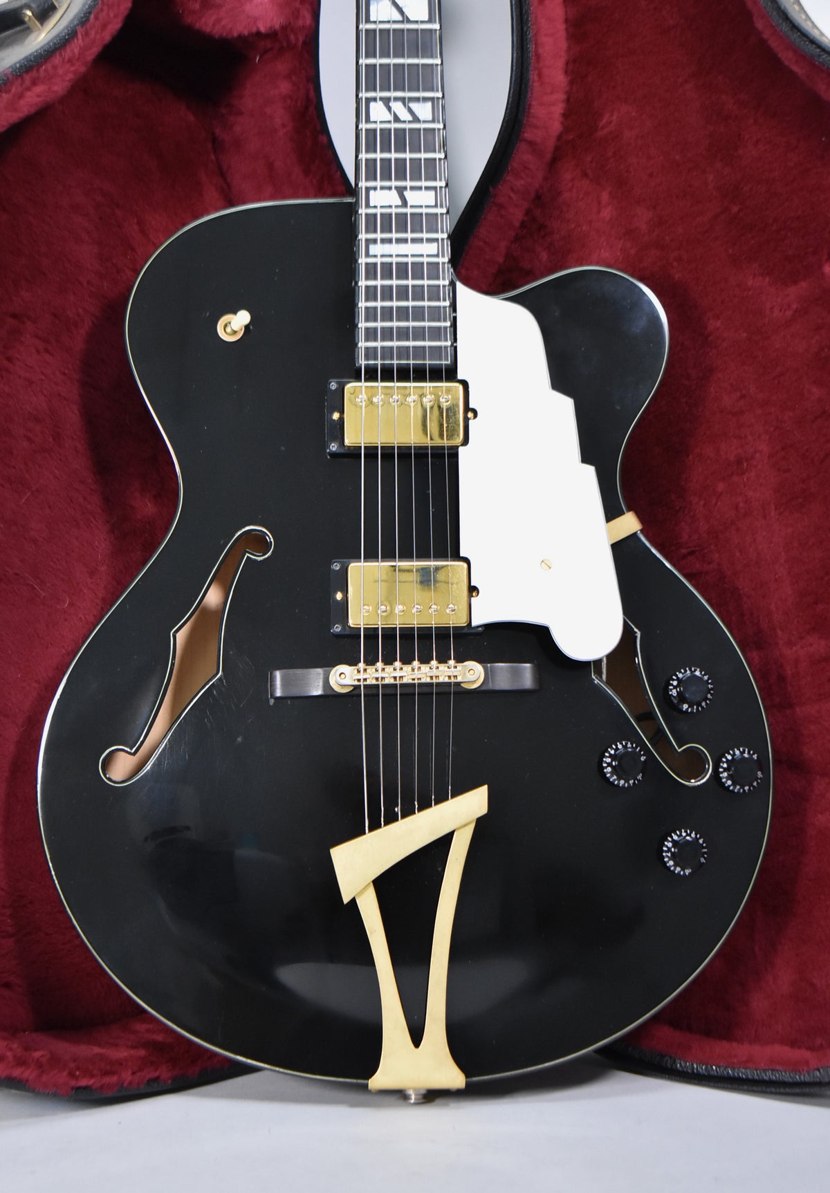 A black archtop electric guitar with gold hardware, f-holes, and a white pickguard, shown in a red velvet case.