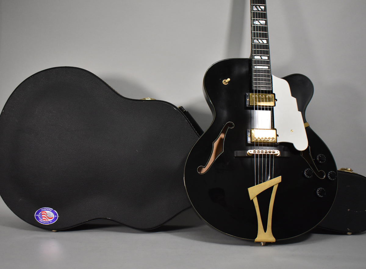 The black archtop guitar, with a white pickguard and gold hardware, stands next to its open black hard case.