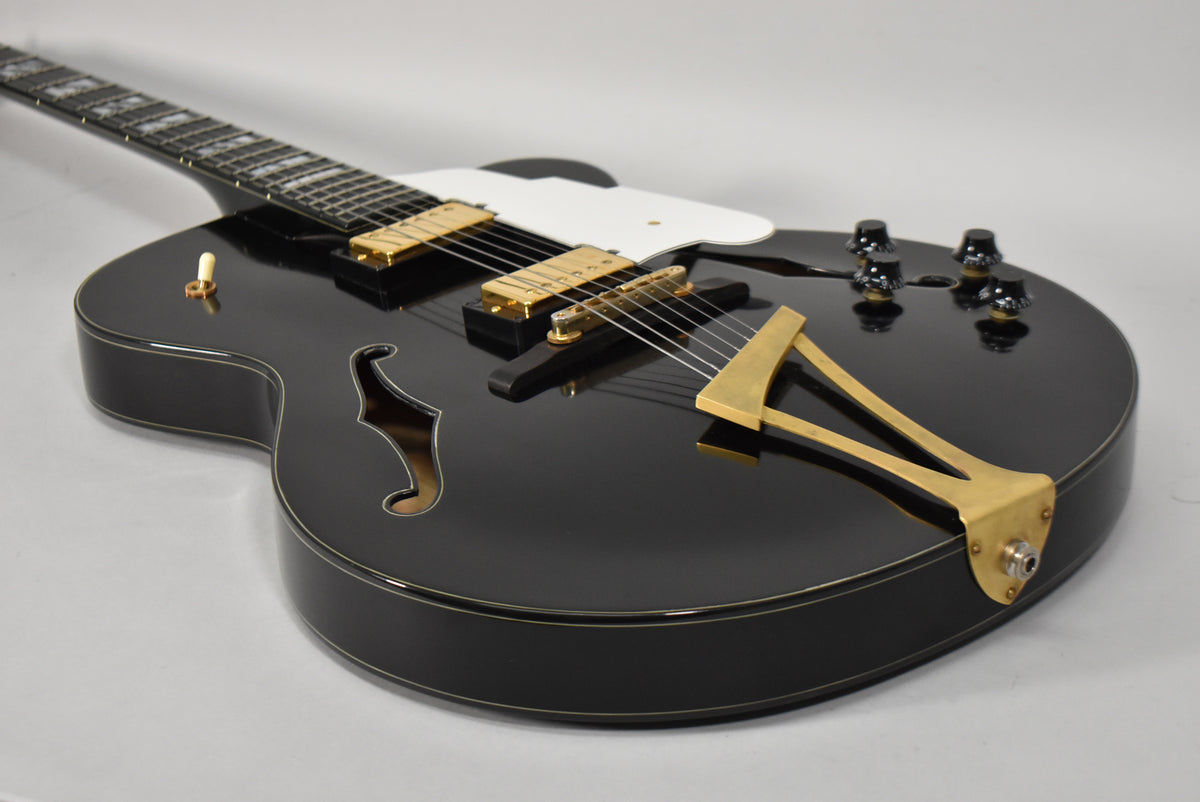Black archtop guitar, angled from the headstock, showing the glossy body, gold hardware, white pickguard, and f-holes.