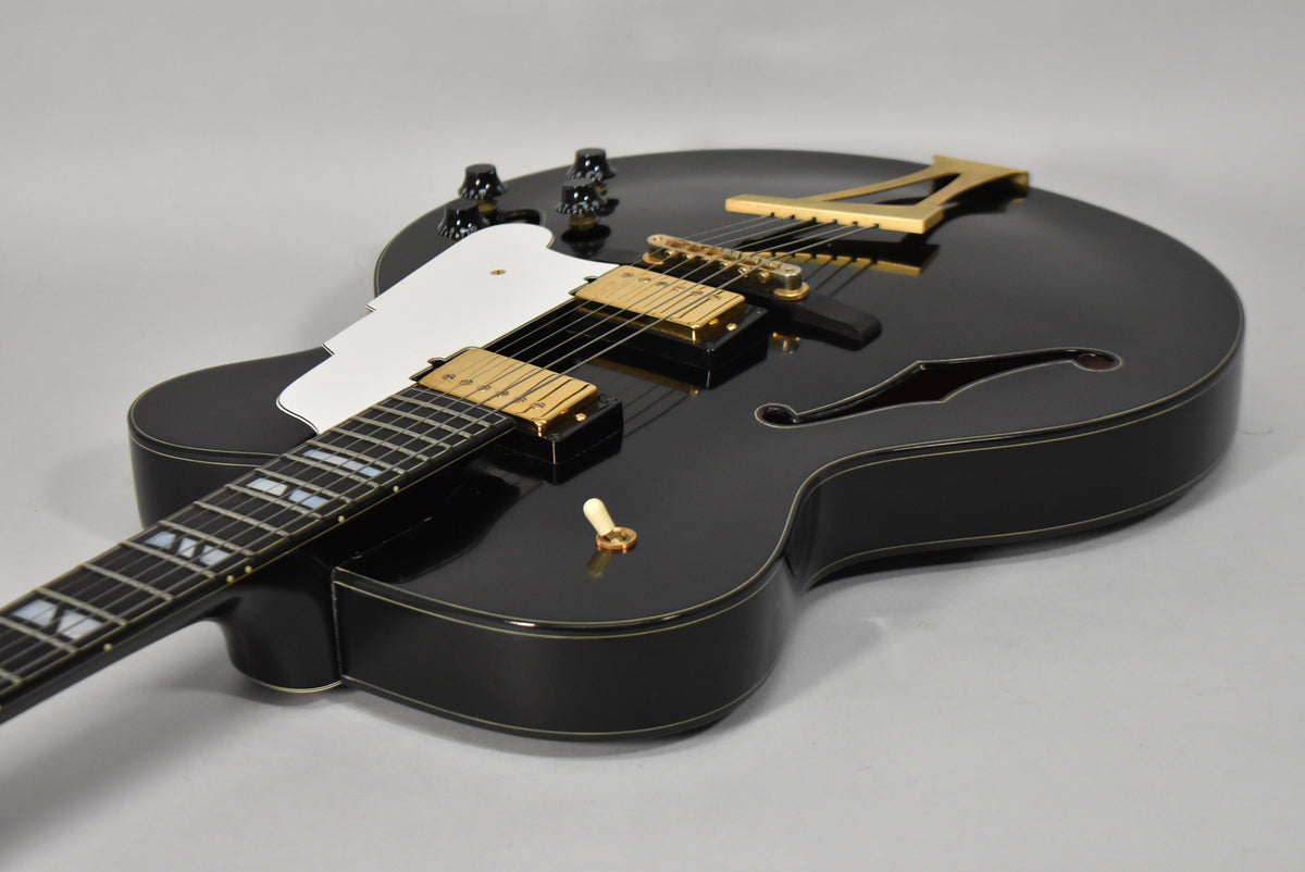 Black archtop electric guitar, angled view of the body and neck, showing gold hardware, white pickguard, and f-holes.