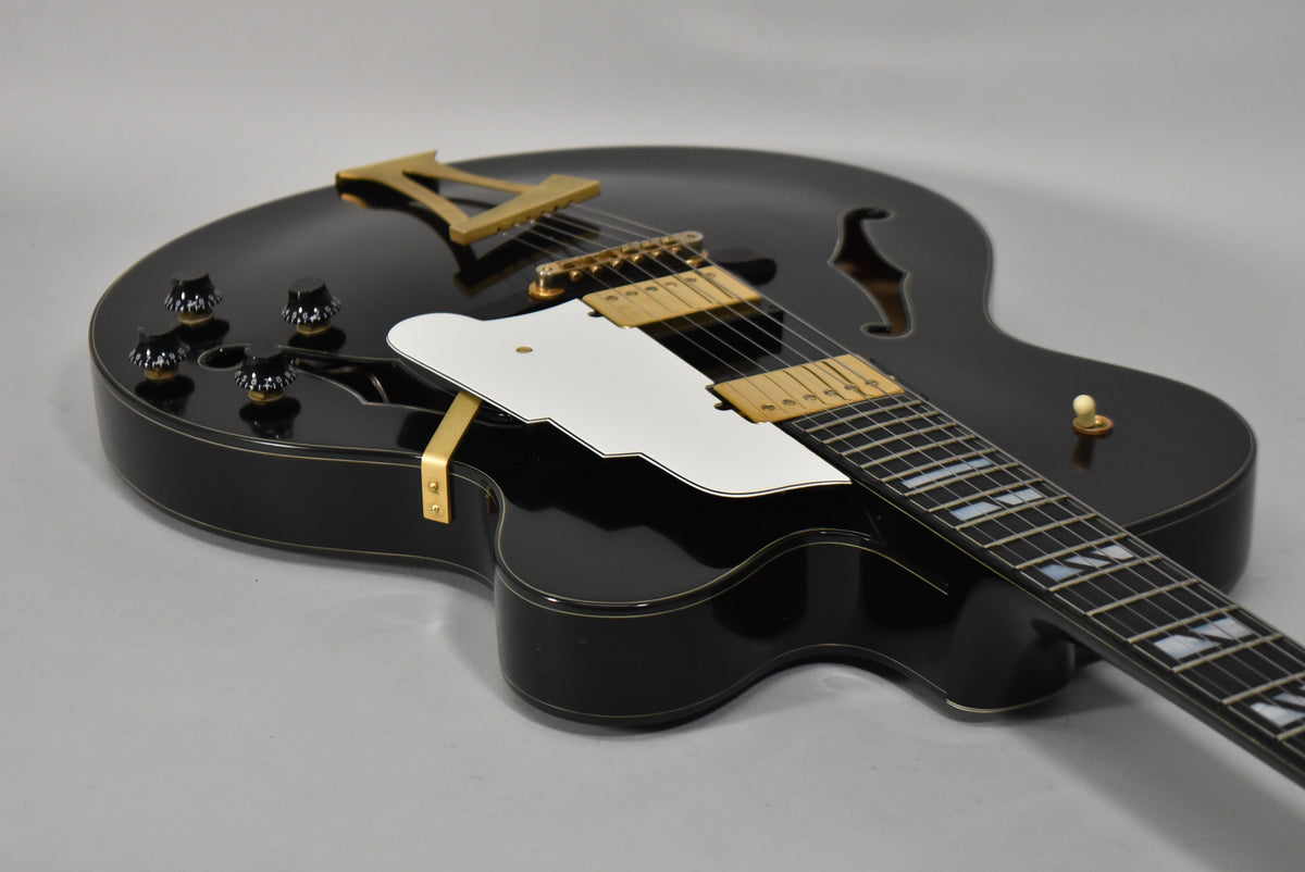 The black archtop guitar is angled from the lower bout, showing gold hardware, a white pickguard, and four black control knobs.