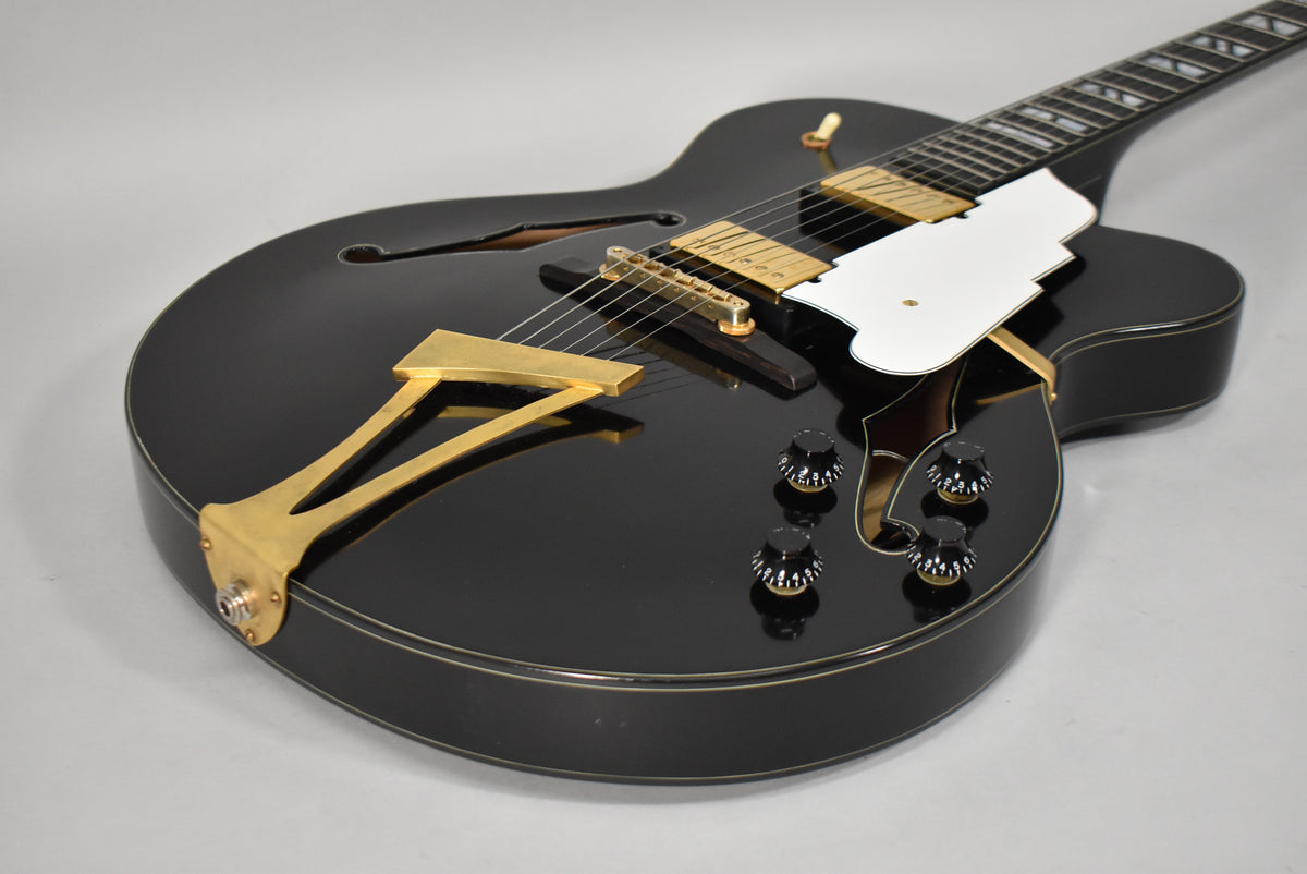 Angled view of a black archtop guitar with gold hardware, including two humbuckers, a trapeze tailpiece, and four control knobs.