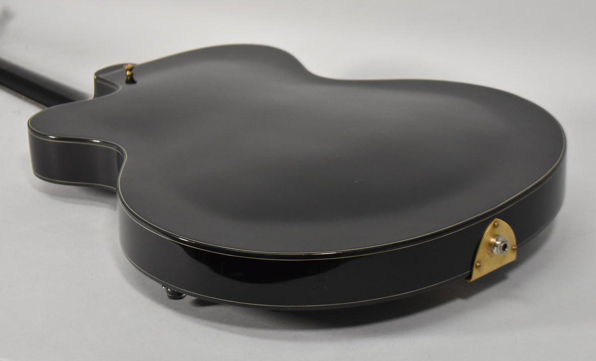 The guitar's glossy black back and sides are shown from a low angle, revealing a gold output jack plate and white binding.
