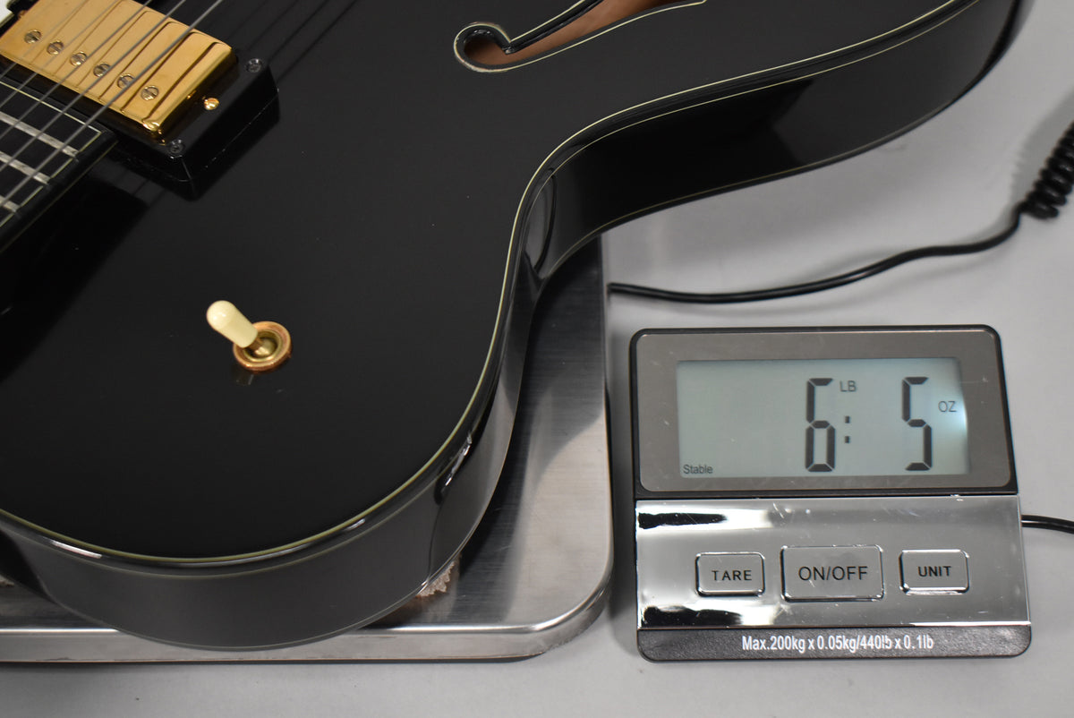 The black archtop guitar's lower bout rests on a scale displaying 6 lbs 5 oz, with a gold pickup and cream switch tip visible.