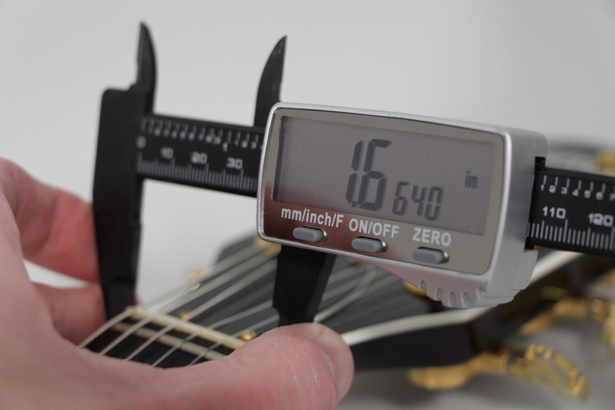 A hand holds a digital caliper measuring the guitar's nut width at 1.640 inches, showing the black headstock and gold tuners.