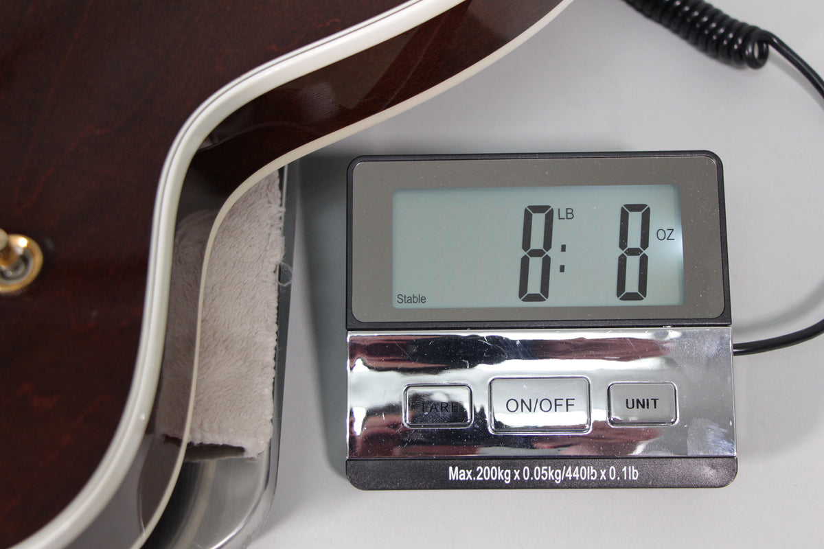 Close-up of the guitar's dark brown body edge on a digital scale displaying 8 pounds, 8 ounces, with a gold strap button visible.