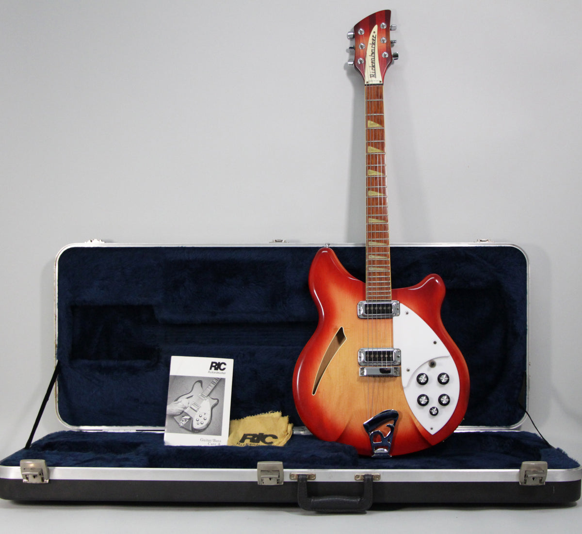 A full-length Fireglo semi-hollow guitar with a white pickguard and five black-capped knobs stands in its open, dark blue plush case.