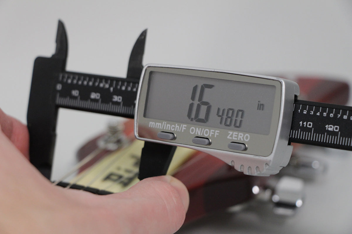 A digital caliper measures the guitar's string height at 1.6mm, with a finger holding the tool against the Fireglo finish.