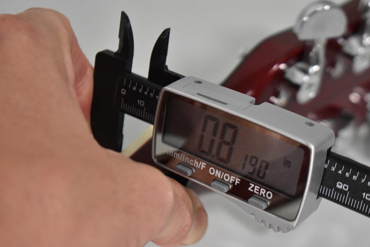 A hand holds a digital caliper measuring the guitar's nut, showing 0.8mm or 0.190 inches, with the cherry headstock blurred behind.