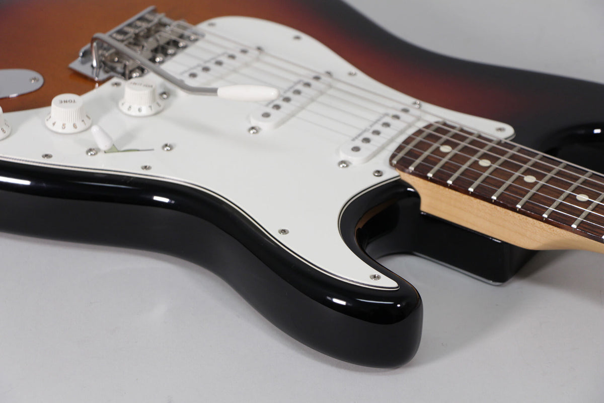 The Stratocaster's lower body and neck are angled from the left, showing the sunburst finish, white pickguard, three single-coil pickups, and rosewood fretboard.