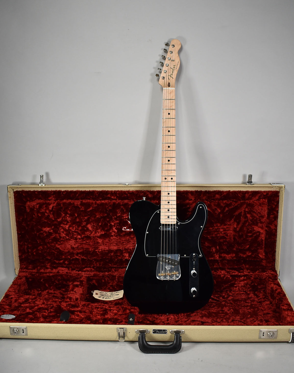 Full-length view of a glossy black Telecaster with a light maple neck, standing upright in its open, cream-colored hard case with red plush lining.