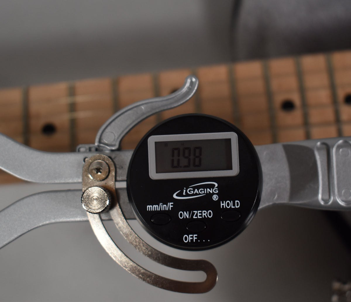 Close-up of a digital fret height gauge showing "0.98" on its screen, clamped to the light-colored maple fretboard.