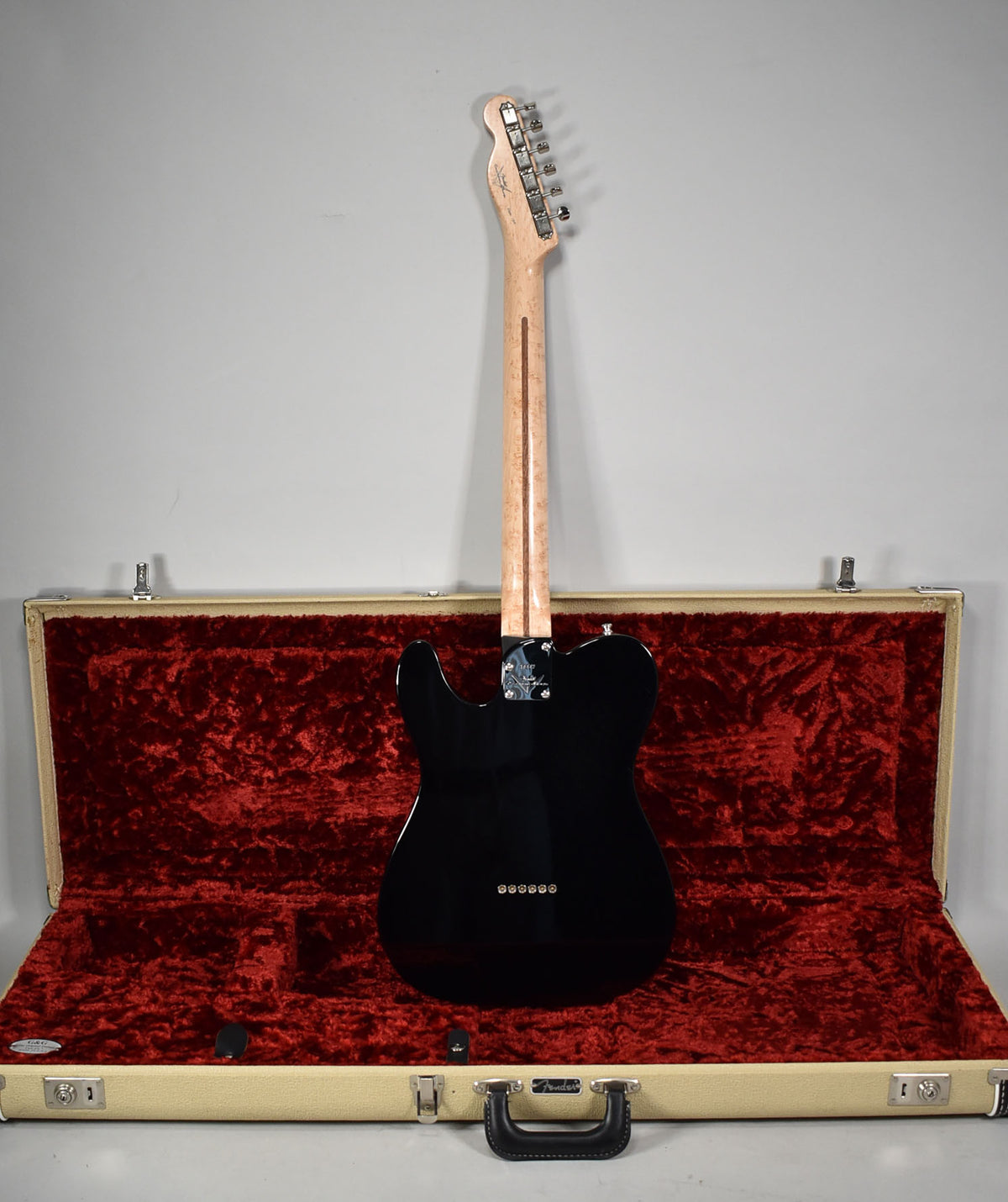 The black Telecaster is viewed from the back, showing its birdseye maple neck and a cream hard case with a red plush interior.