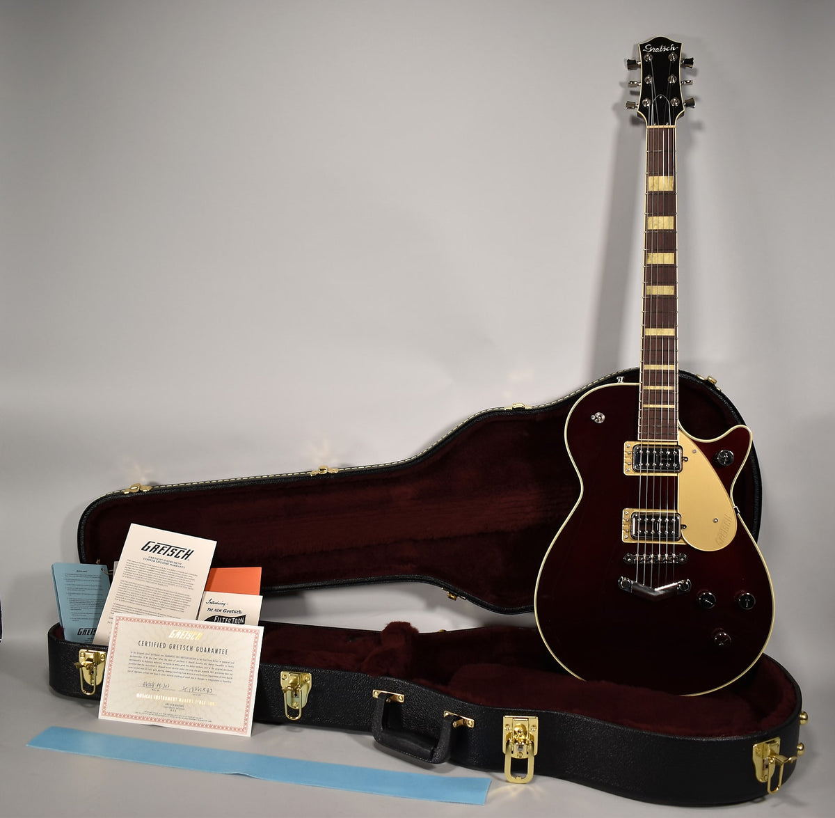 Full view of a dark red electric guitar with a cream pickguard and gold hardware, standing upright in its open hard case.