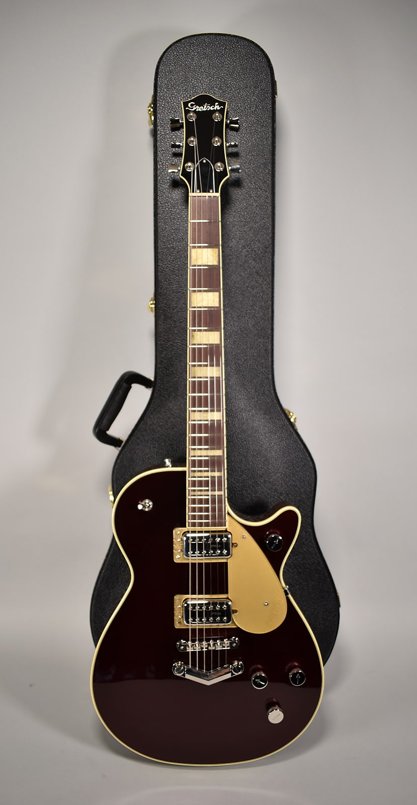 Full-length view of a dark red electric guitar with a gold pickguard, gold humbuckers, and block inlays, standing in a black hard case.