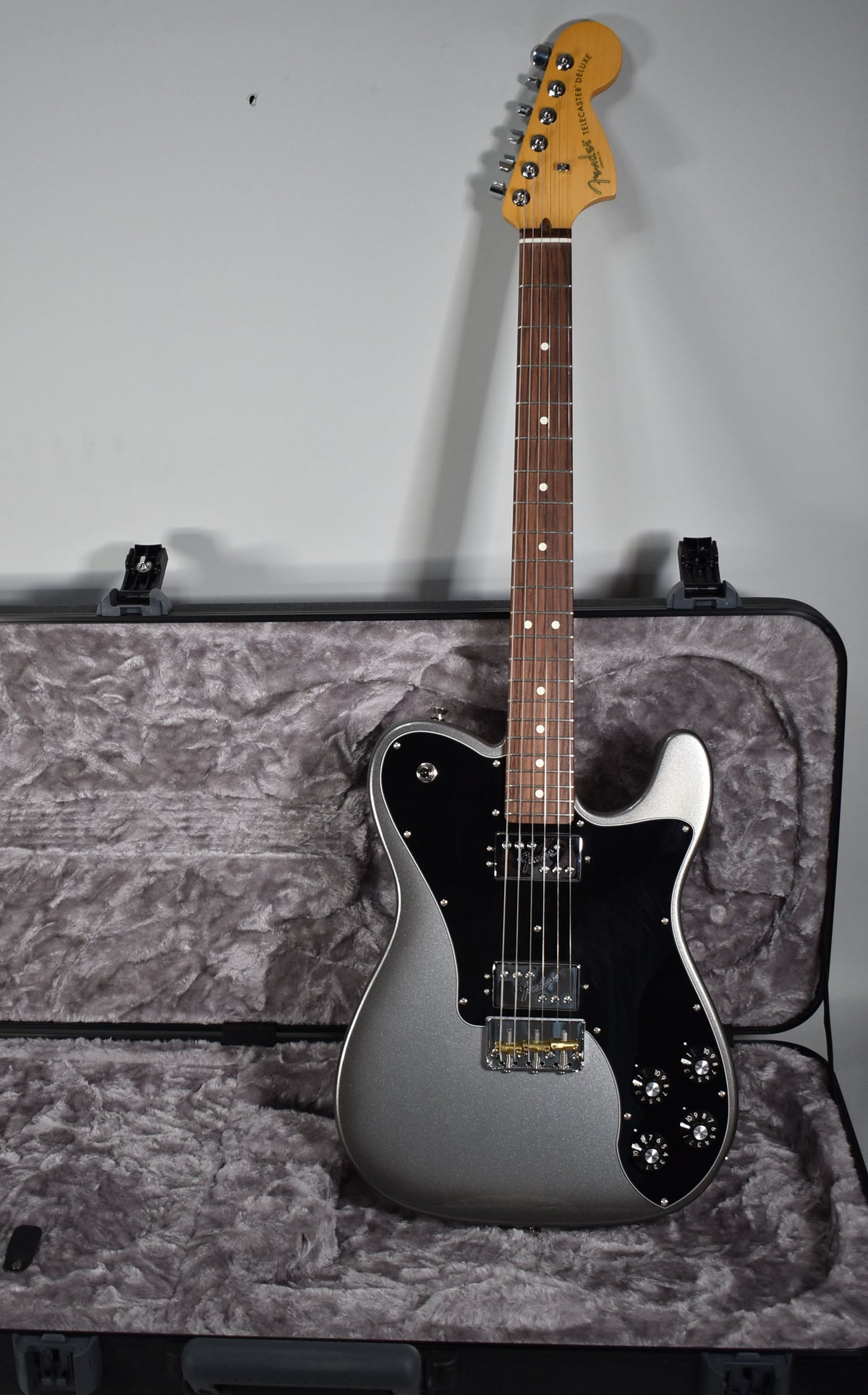 A silver Telecaster Deluxe with a black pickguard and a rosewood fretboard rests in a black hard case with gray plush lining.