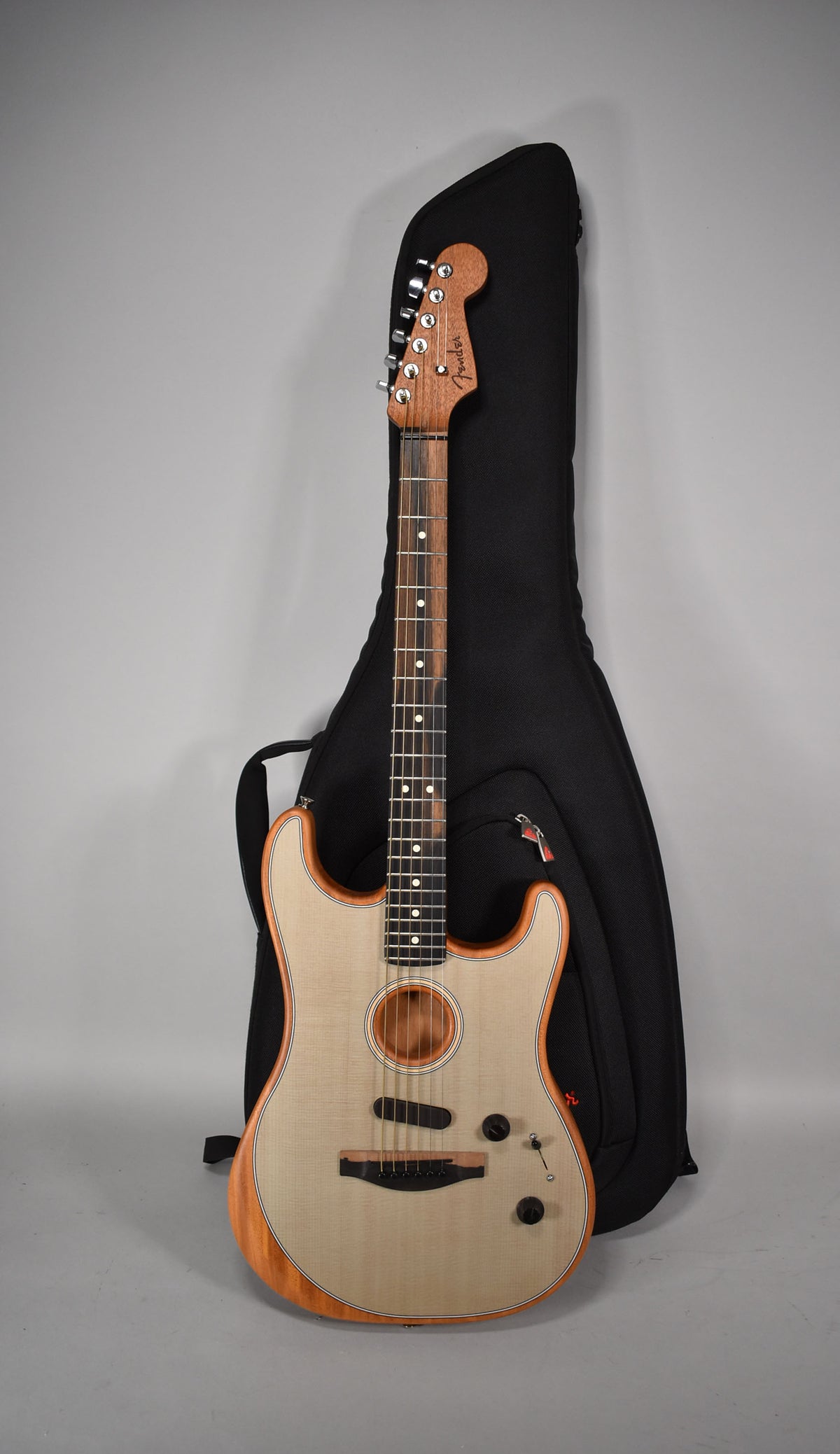 Acoustasonic Stratocaster with a light wood top, dark wood sides, neck, and headstock, standing in front of a black gig bag.