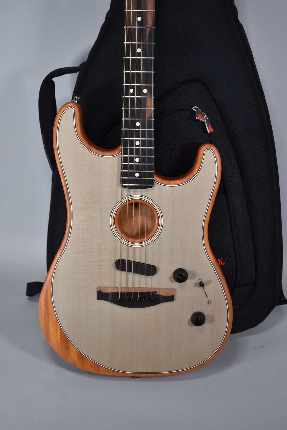 A light wood acoustic-electric guitar with a dark fretboard and a black gig bag behind it.