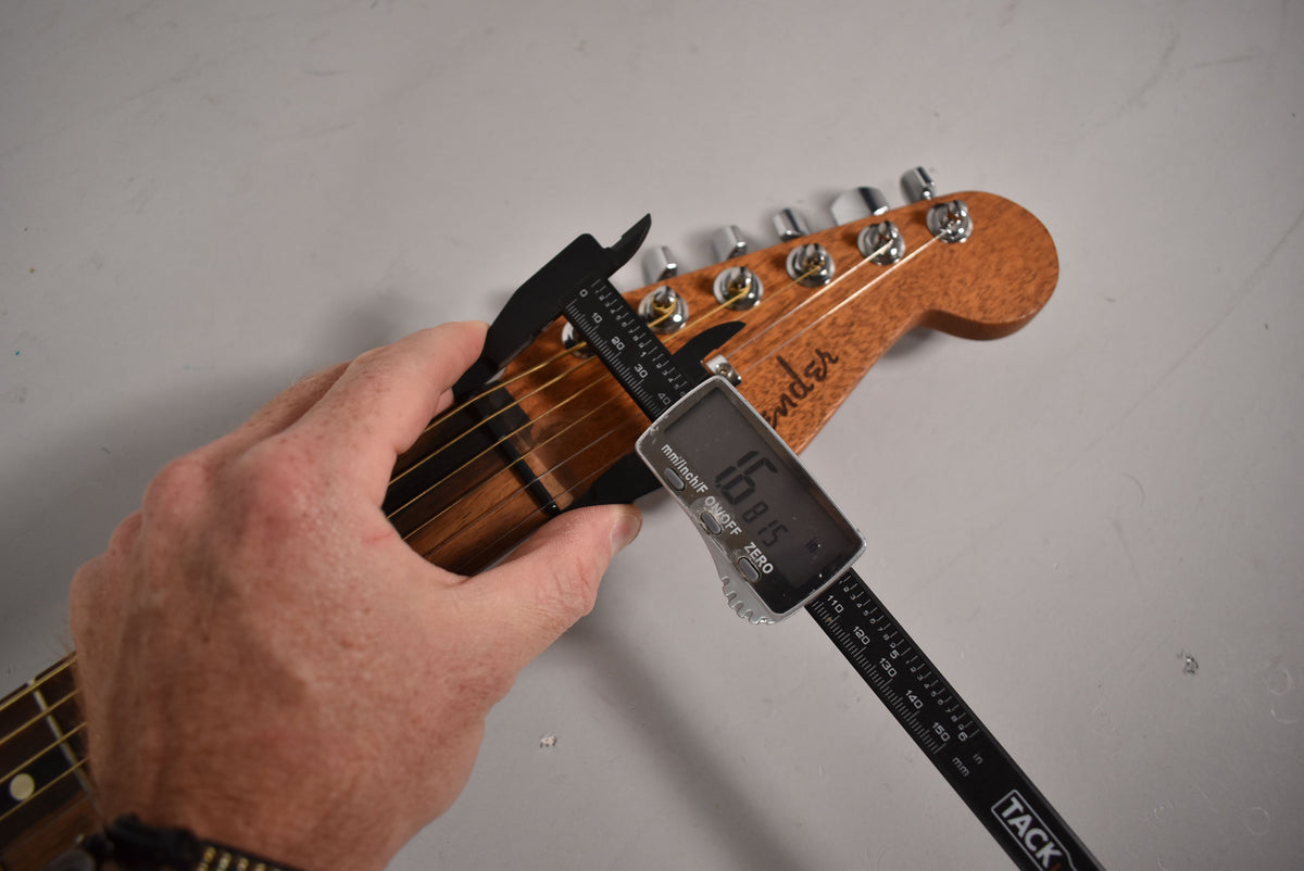A hand holds a digital caliper measuring the nut width of the guitar's light brown headstock, showing 1.6815 inches.