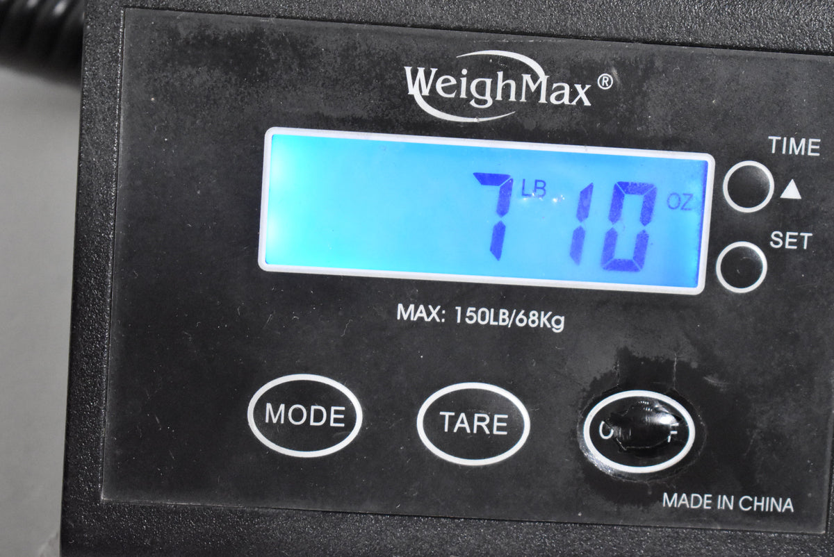 Close-up of a black digital scale displaying "7 LB 10 OZ" in bright blue, with "WeighMax" and "MADE IN CHINA" visible.