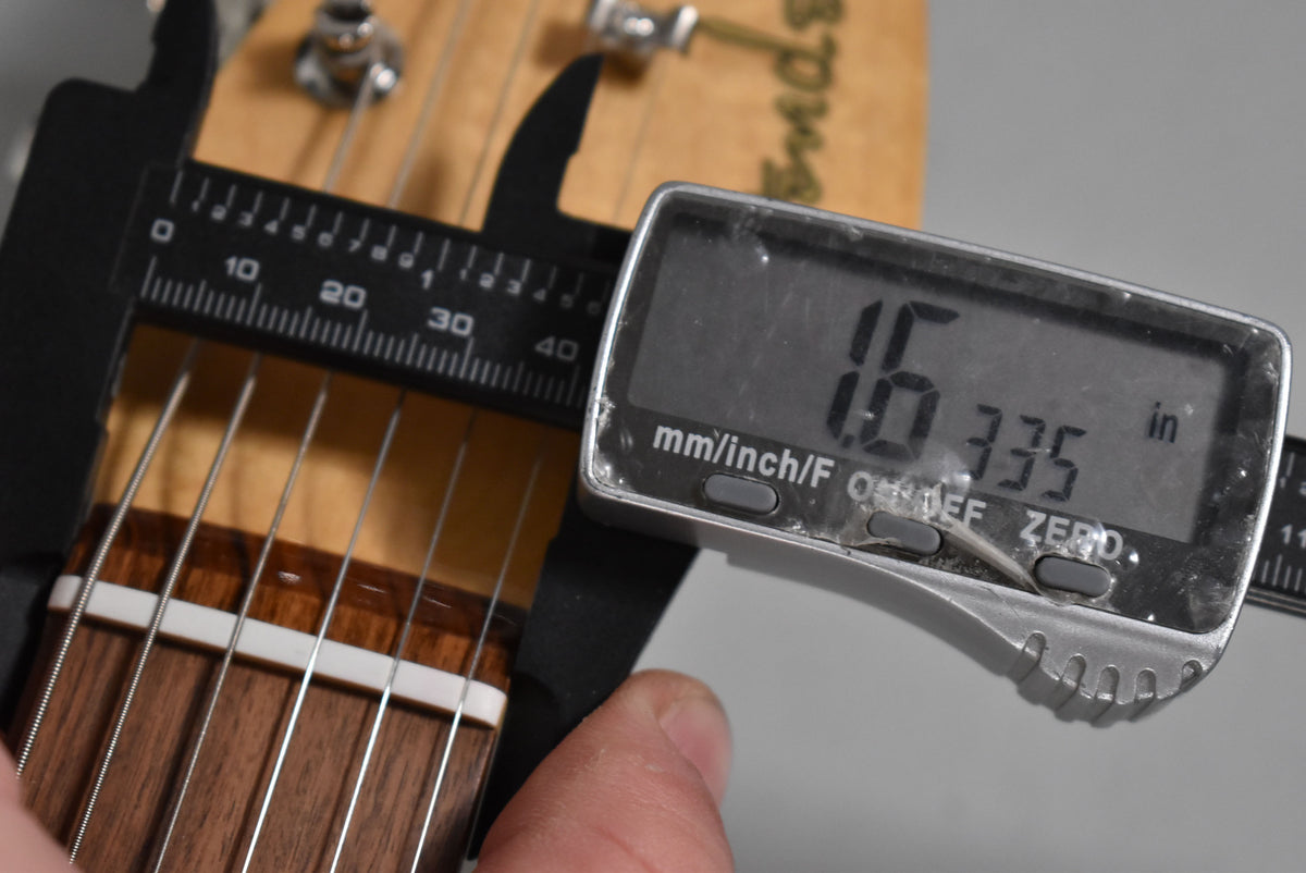 A digital caliper measures the nut width of the guitar's dark wood fretboard, showing 1.6335 inches on its display.