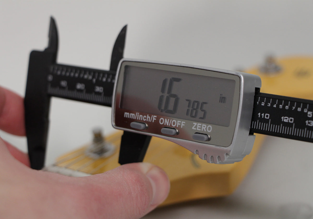 A digital caliper measures the nut width of a light-colored guitar neck, displaying "1.6785 in" on its screen.